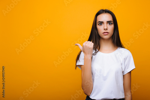 Beautiful brunette pointing confused away with finger dressed in white shirt against yellow wall.