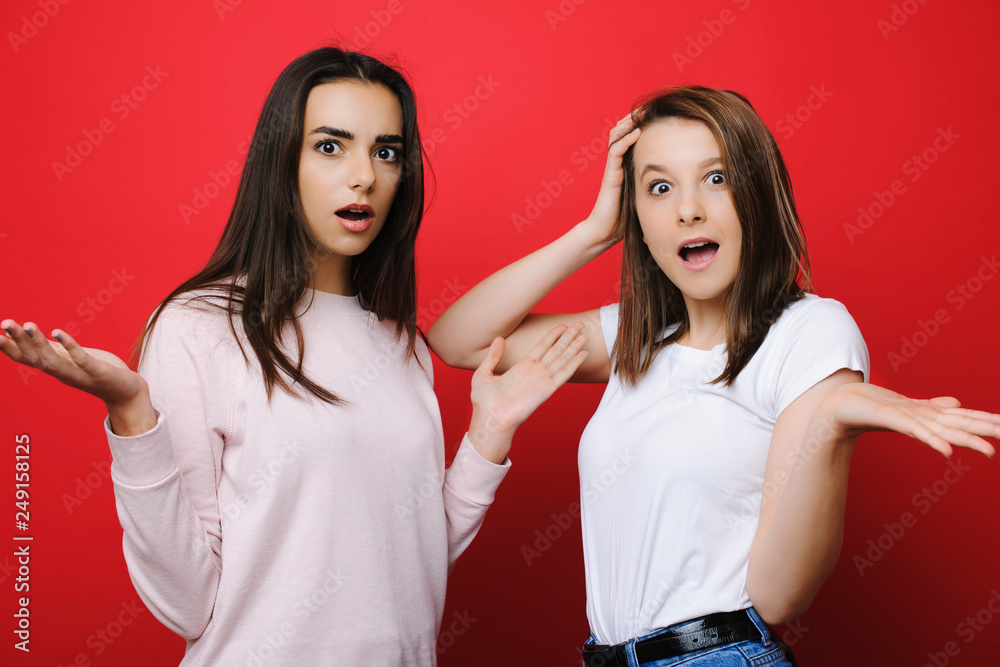 Two amazing girl friends looking into camera amazed with open mouths gesticulates with their hands on a red background. Two amazing girl friends looking into camera amazed with open mouths gesticulates with their hands on a red background.