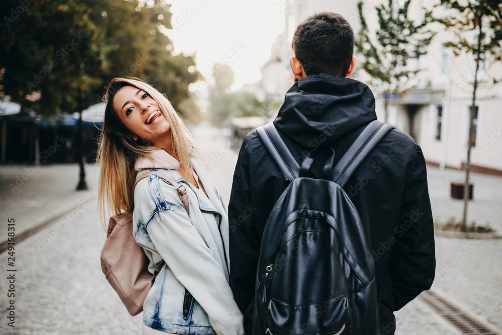 Amazing couple wearing backpacks traveling while girl is looking into camera and showing her tongue smiling.