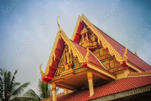 Colorful gable of the public Buddhist church in the rural area of Thailand that the church building decorated with red tiles roof under blue sky and white clouds on background.