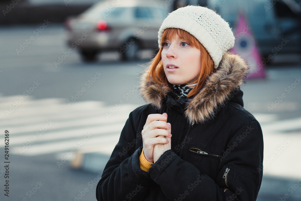 catalogus bitter tragedie red-haired woman wearing white knitted hat and dressed in warm hooded  casual parka jacket outerwear, girl in the parking lot, pedestrian crossing  and car in the background, girl warms frozen hands Stock