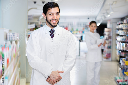 Portrait of male specialist who is standing near shelves with medicines in pharmacy.