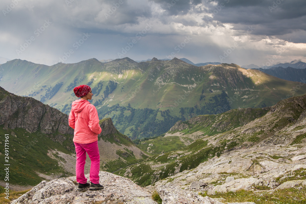 view from the back on girl in pink pants standing on a cliff  hands in pockets on a mountain background under storm sky