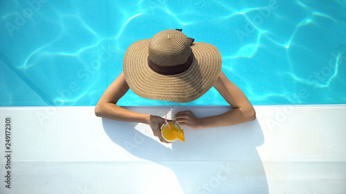 Young woman in elegant hat swimming in pool and enjoying delicious cocktail