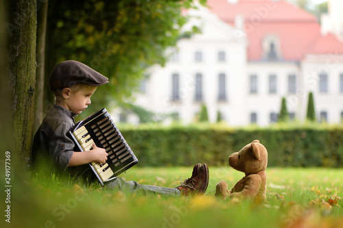 Fotografie A four years old boy sitting on grass and play accordion for teddy bear