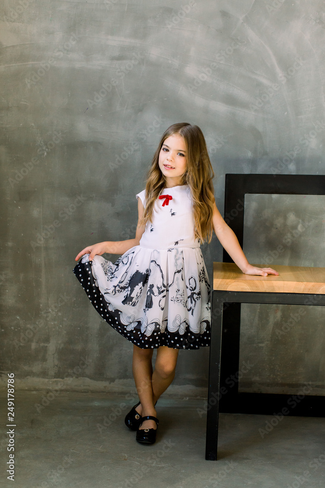 Naklejka premium A portrait of a full-height little gir that put a hand on a chair on a gray background