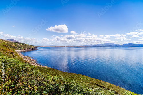 Blue sky with white puffy clouds over rocky costal line and mountains in a distance.