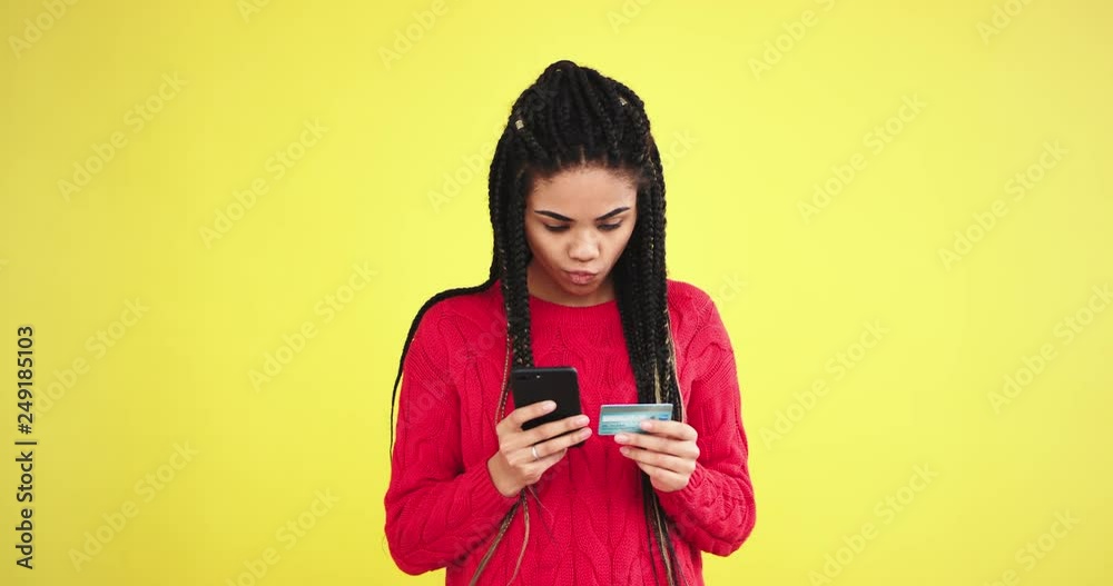In a studio with yellow background wall african lady in a red sweater in front of the camera using a smartphone and bank card to order something In a studio with yellow background wall african lady in a red sweater in front of the camera using a smartphone and bank card to order something