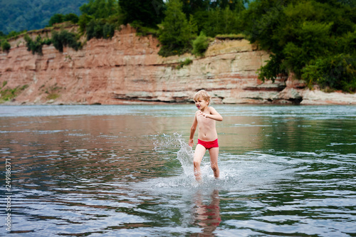 Blonde smiling boy in red swimsuit running in the mountain river