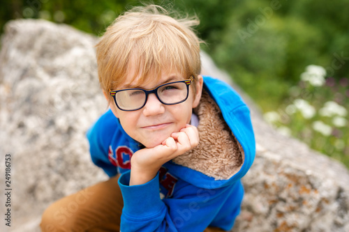 Nice funny blonde boy with strabismus wearing glasses with special lens sits on the stone in the mountains photo