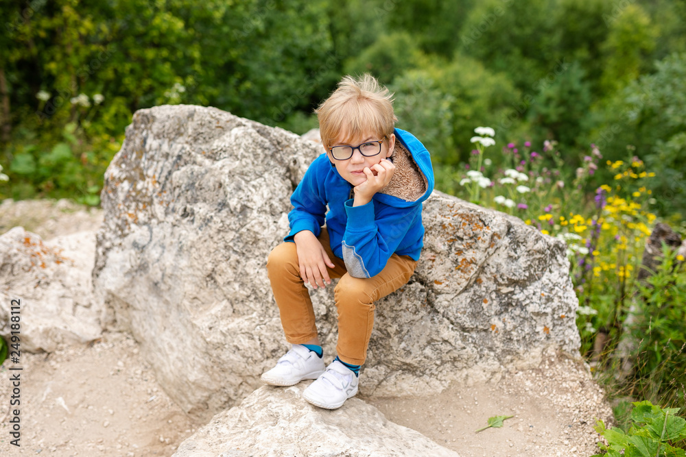 Nice funny blonde boy with strabismus wearing glasses with special lens sits on the stone in the mountains Nice funny blonde boy with strabismus wearing glasses with special lens sits on the stone in the mountains