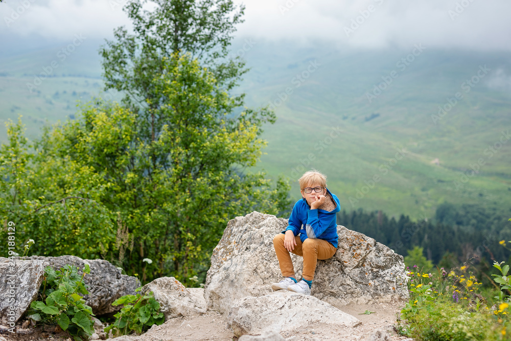 Naklejka premium Nice funny blonde boy with strabismus wearing glasses with special lens sits on the stone in the mountains