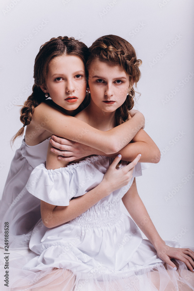 Two teenage fashionable girls models sisters hugging together in white dresses on isolated grey background. Fashion, glamourous, friendship concept Two teenage fashionable girls models sisters hugging together in white dresses on isolated grey background. Fashion, glamourous, friendship concept