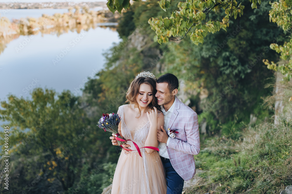 Autumn portrait of beautiful newlyweds, on the background of green trees and rocks with bushes. Wedding photography. Stylish groom hugging a young bride in a beige dress with a crown.