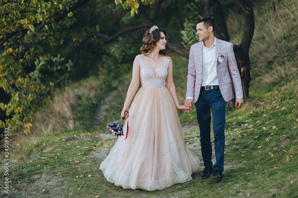 Autumn portrait of newlyweds in love, on a background of green trees on nature. Wedding photography. Stylish groom in a plaid jacket walking with a cute bride in a beige dress with a crown. Autumn portrait of newlyweds in love, on a background of green trees on nature. Wedding photography. Stylish groom in a plaid jacket walking with a cute bride in a beige dress with a crown.