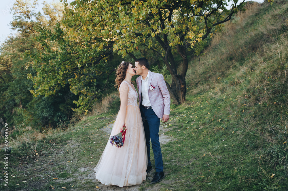 Autumn portrait of newlyweds in love, on a background of green trees on nature. Wedding photography. Stylish groom in a plaid jacket hugging a pretty bride in a beige dress with a crown. Autumn portrait of newlyweds in love, on a background of green trees on nature. Wedding photography. Stylish groom in a plaid jacket hugging a pretty bride in a beige dress with a crown.