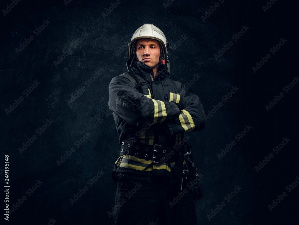 Portrait of a fireman in uniform and helmet stands with crossed hands, looking sideways with a confident look. Studio photo against a dark textured wall