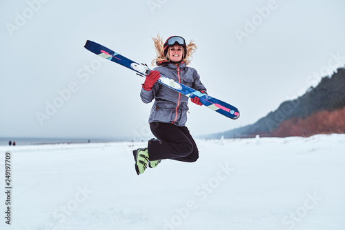 Cheerful woman wearing warming sportswear holding skis and jumping on a snowy beach
