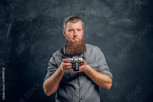A man with a stylish haircut and beard wearing a gray shirt holding a photo camera. Studio shot on a gray textured wall