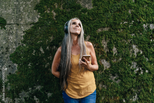 Woman with cell phone standing in front of wall listening music with headphones photo