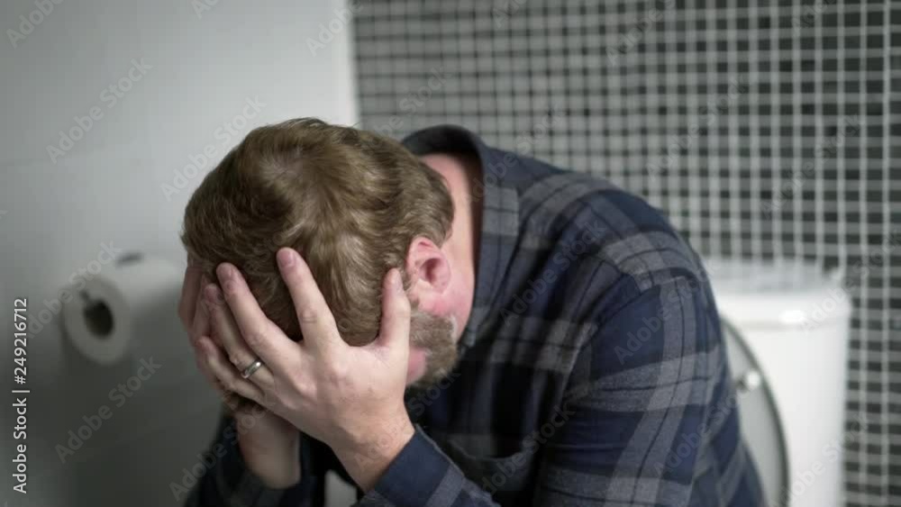 Constipated man sits on toilet with his head in hands, suffering in