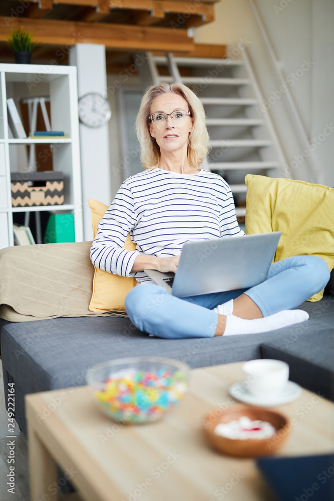 Full length portrait of contemporary mature woman looking t camera while using laptop sitting on sofa Full length portrait of contemporary mature woman looking t camera while using laptop sitting on sofa