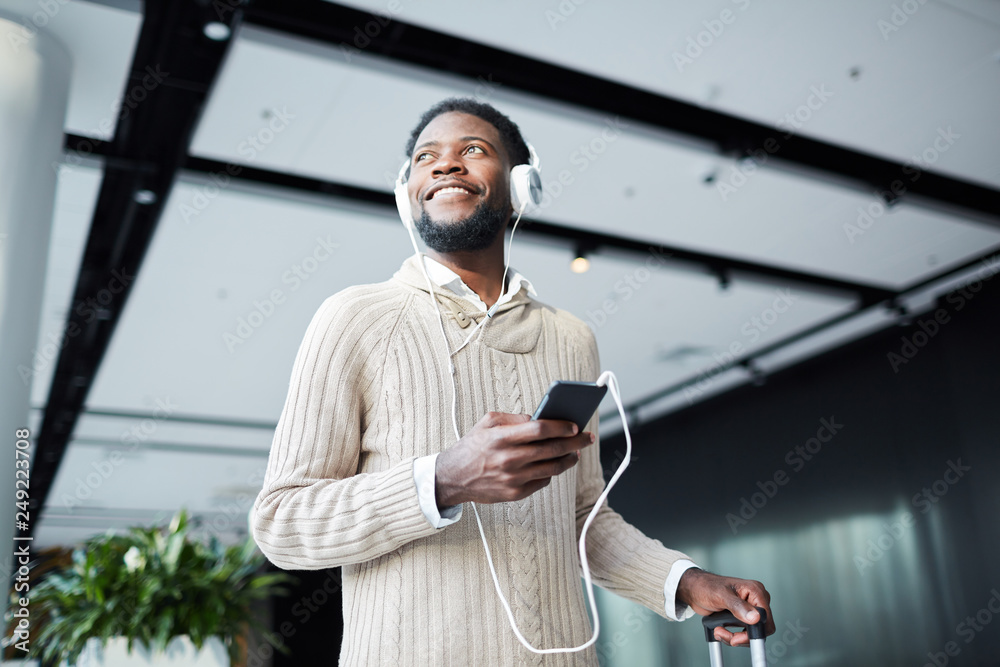 Happy young casual traveler with smartphone and headphones listening to his favorite music in airport lounge Happy young casual traveler with smartphone and headphones listening to his favorite music in airport lounge