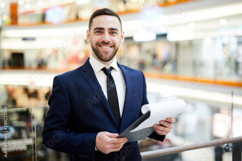 Young cheerful businessman with financial document standing in front of camera in the mall