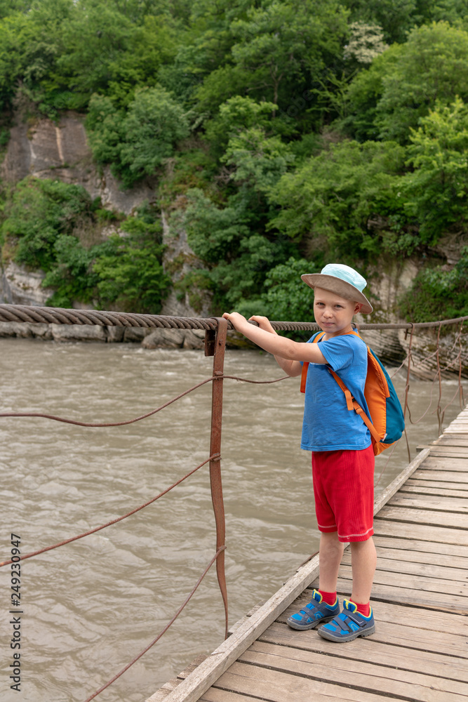 A boy with a backpack crosses a mountain river with rocky shores on a suspension bridge.