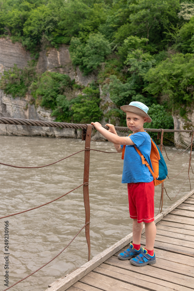 A boy with a backpack crosses a mountain river with rocky shores on a suspension bridge.