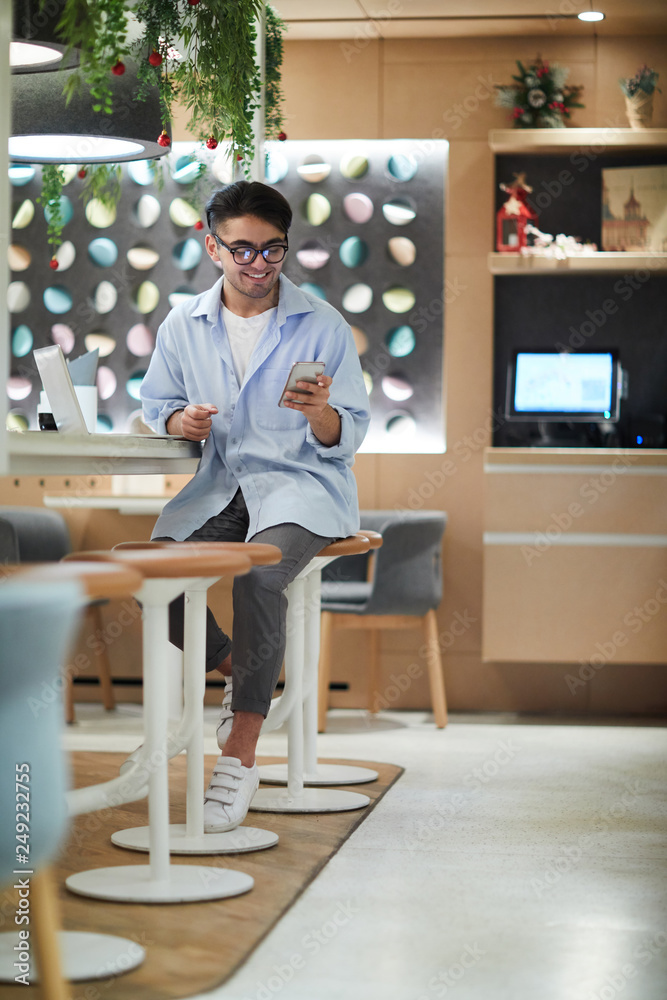 Happy casual guy sitting in college cafe, waiting for his friends and scrolling in smartphone Happy casual guy sitting in college cafe, waiting for his friends and scrolling in smartphone