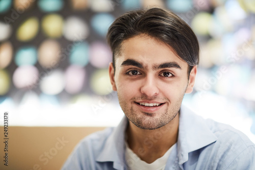 Young smiling handsome man in light blue shirt looking at you while sitting in front of camera in cafe