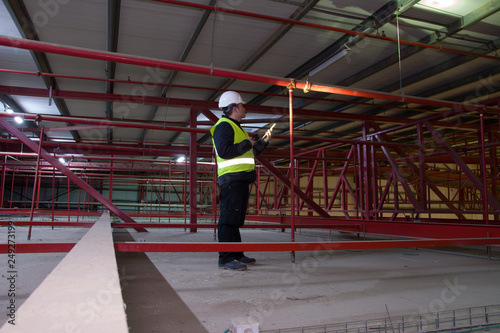 Technician engineer in a whute helmet checks the sprinklers pipes and roof constractions with a tablet.