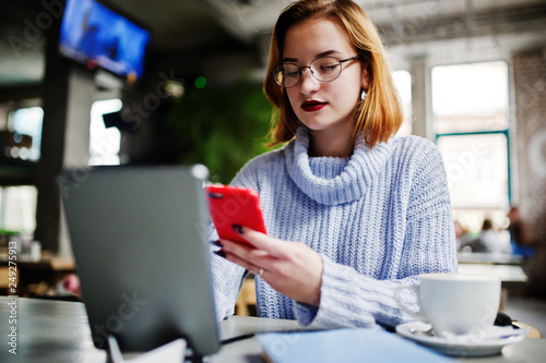 Cheerful young beautiful redhaired woman in glasses using her phone, touchpad and notebook while sitting at her working place on cafe with cup of coffee.