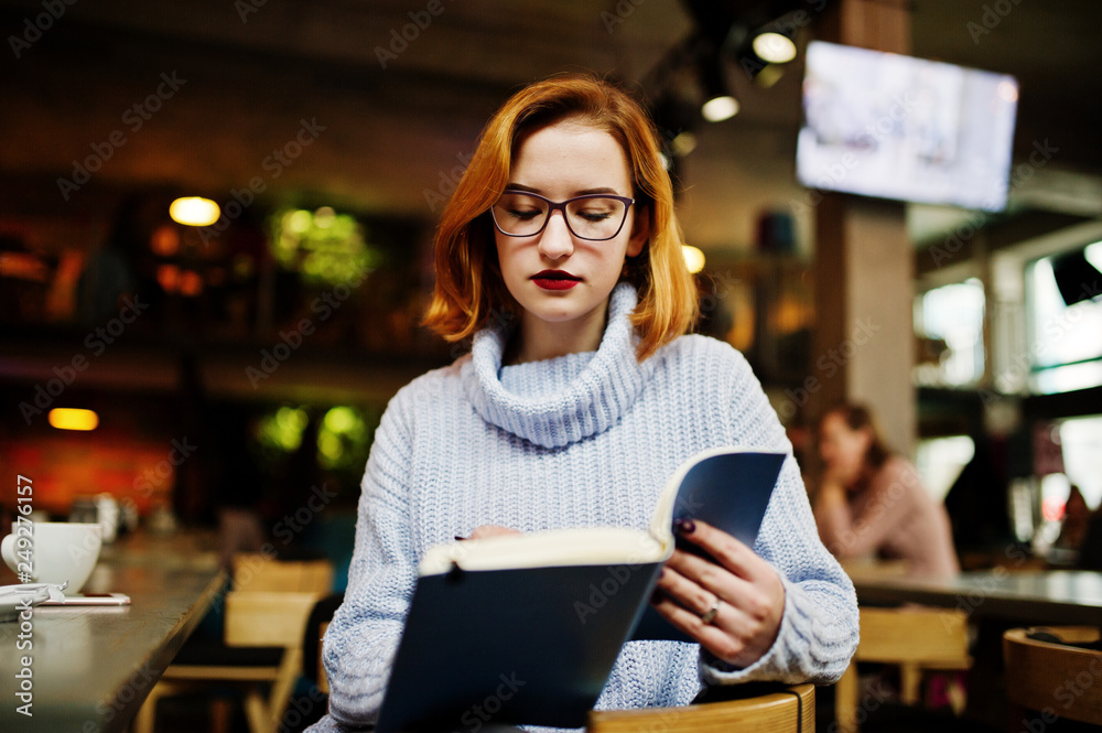 Cheerful young beautiful redhaired woman in glasses sitting at her working place on cafe reading something at her notebook. Cheerful young beautiful redhaired woman in glasses sitting at her working place on cafe reading something at her notebook.