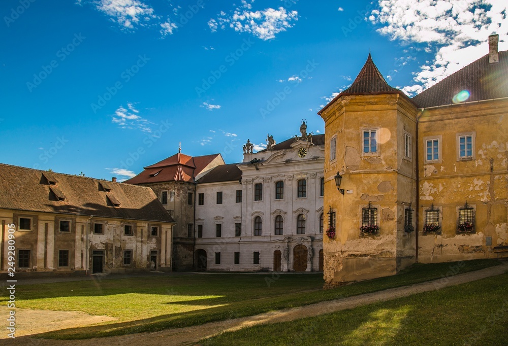 Kladruby, Czech Republic / Europe - July 7 2018: Monastery of Benedictines from 12th century, white and yellow buildings, wall clock, green grass, sunny summer day, sun beam, blue sky with clouds
