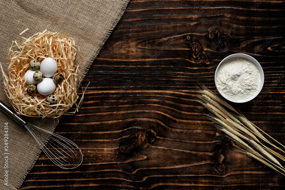 Flour in a white bowl, dark wood background with ears of wheat, eggs in straw nest, top view with copy space. ingredients for bakery products