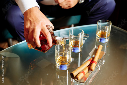 man pours alcohol in three glasses, three cigars lie in an ashtray
