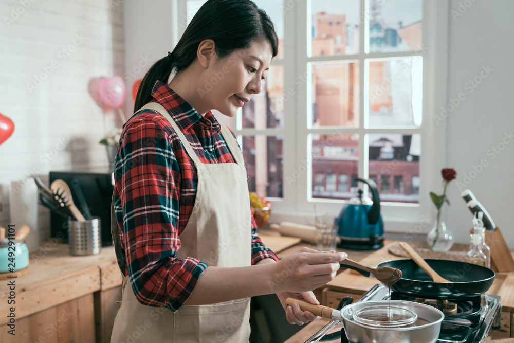 young girl in apron holding wooden spoon stirring soft melted rich chocolate in hot pot on stove. elegant asian woman cooking handmade sweet cocoa dessert for valentine day gift in wooden kitchen.