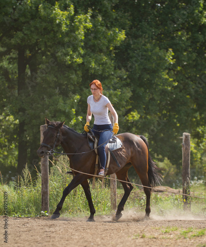 A redhead woman in white t-shirt and glasses riding a horse in nature.