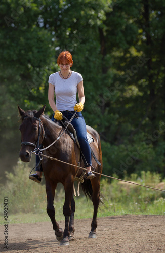 A redhead woman in white t-shirt and yellow gloves riding a horse in forest