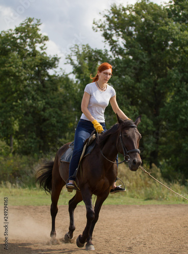 A redhead woman in white t-shirt riding a dark brown horse in nature. © KONSTANTIN SHISHKIN
