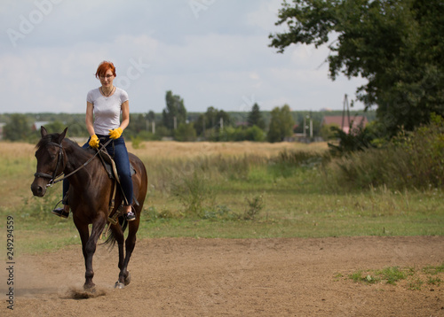 A redhead woman riding a dark brown horse in nature