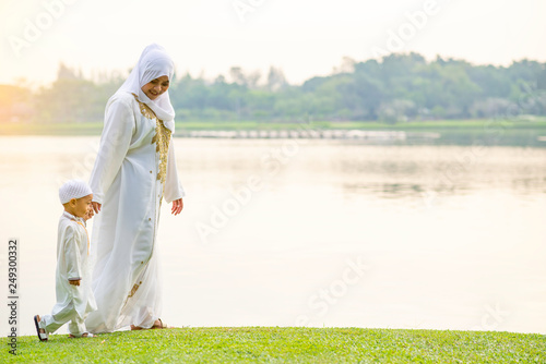Muslim mother with her son they are walking together on grass field near beautiful lake. Muslim family concept.