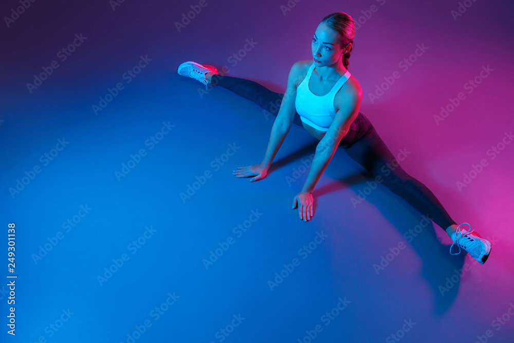 Attractive young slim gymnast woman in sports clothing stretching on the floor in neon lights. Flexible muscular woman doing gymnastic and yoga split.