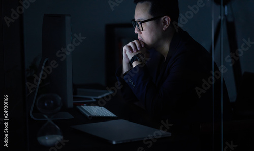 Young Asian businessman working on a laptop while watching computer at his office desk at night .