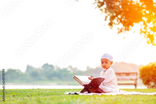 Little Muslim boy reading holy Quran on grass field near beautiful lake.