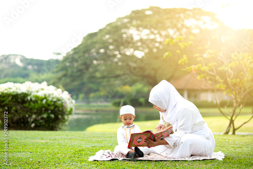 Muslim mother teaching her little son to reading the Quran on grass field near beautiful lake. Muslim family concept. 
