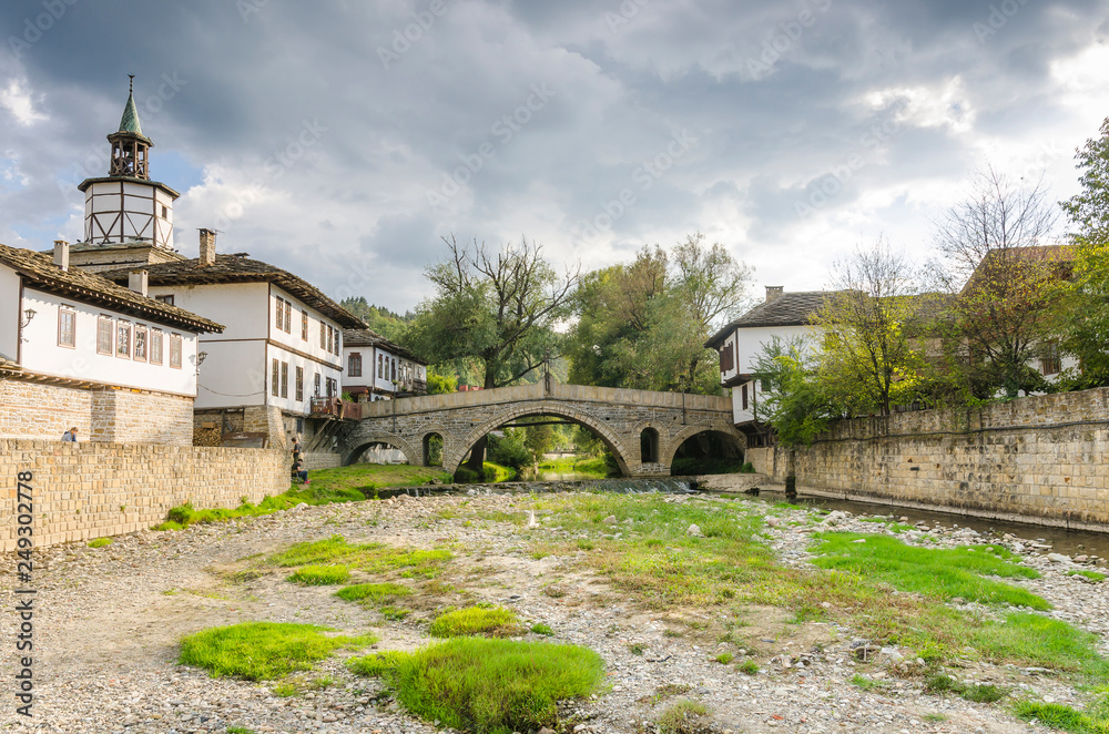 Old bridge (also called hunchback bridge) on the Trevnenska river and the clock tower in town of Tryavna
