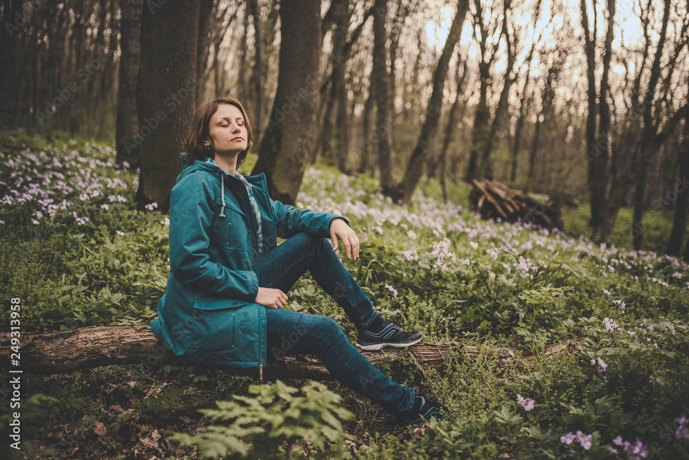 Toned photo of happy hiking middle-aged woman in blue jacket sitting and resting in evening spring forest Toned photo of happy hiking middle-aged woman in blue jacket sitting and resting in evening spring forest